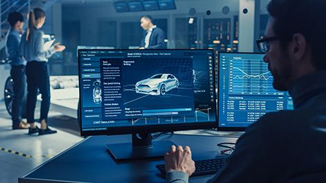 Professional Engineer Works on a Computer with a 3D CAD Software and Tests the Electric Car Chassis Prototype with Wheels, Batteries and Engine Standing in a High Tech Development Laboratory.